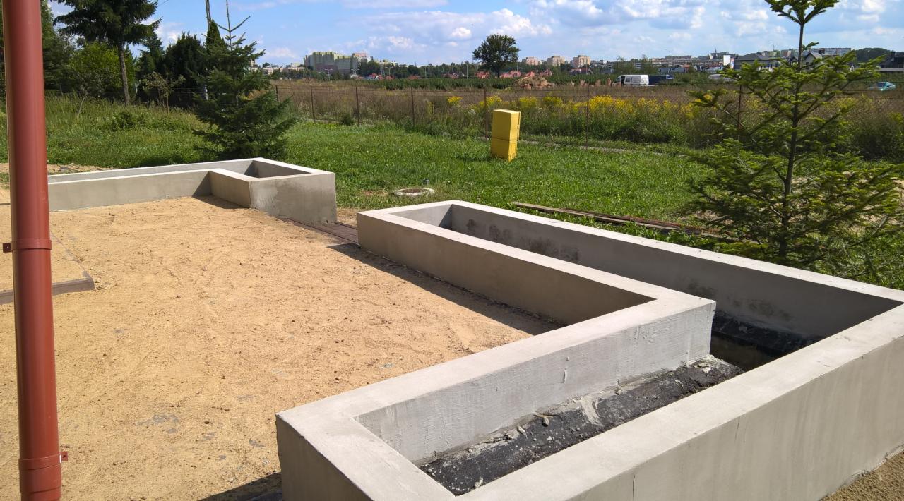 Przygotowanie terenu pod taras: betonowe obramowanie donic w kształcie litery L, wypełnione piaskiem, z widocznym systemem izolacji i odprowadzania wody, w tle trawa i zabudowania.