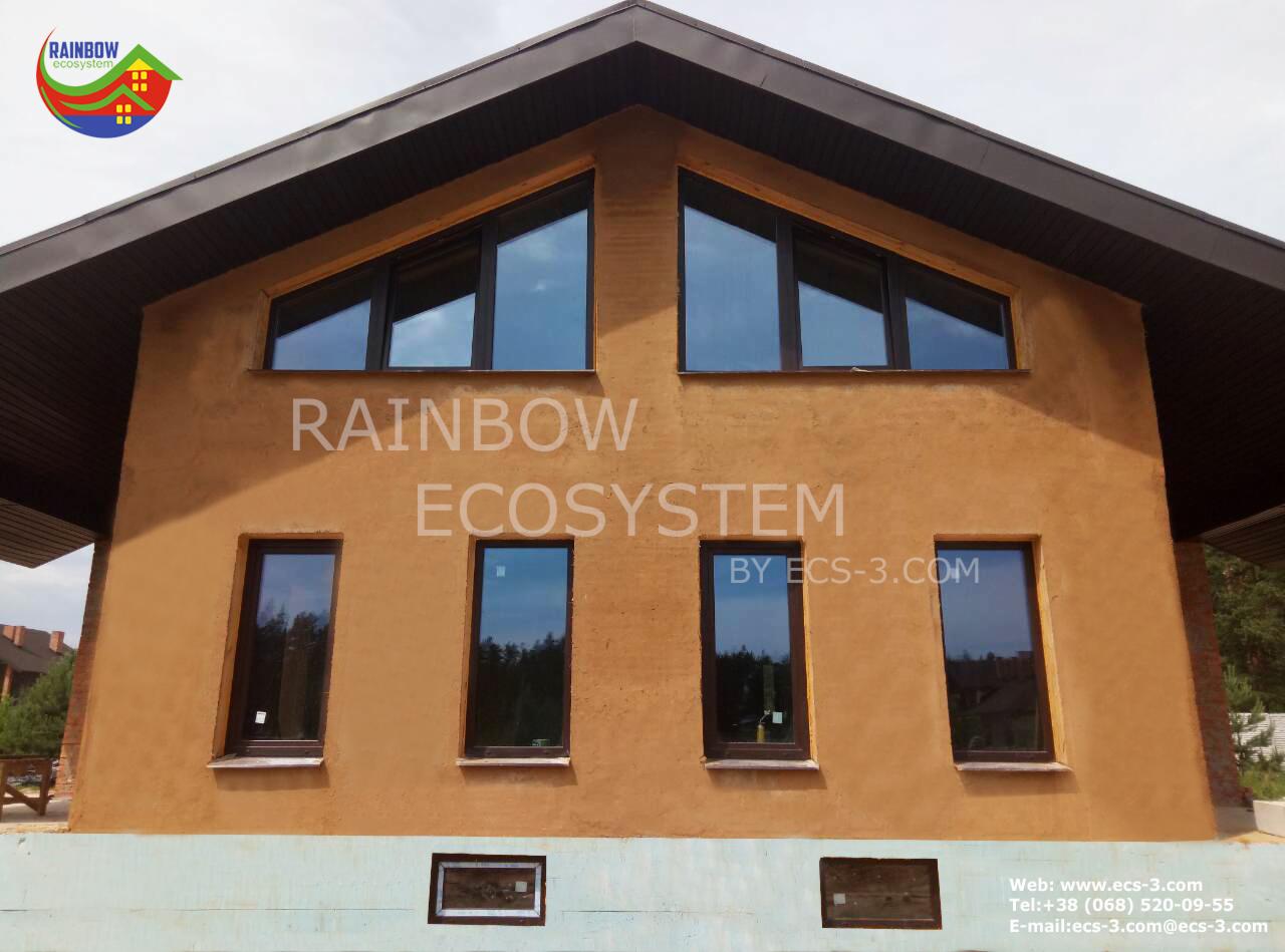 Elewacja budynku z oknami o nietypowych kształtach, pokryta tynkiem w odcieniach beżu i brązu, z widocznym logo firmy 'Rainbow Ecosystem' na ścianie.