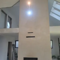 Ściana kominkowa w trakcie tynkowania z widocznymi otworami wentylacyjnymi i wkładem kominkowym, surowy stan wykończenia wnętrza z drabiną w tle.