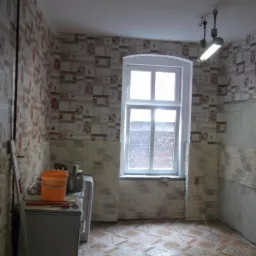 Kuchnia w trakcie remontu: położone płytki imitujące cegłę do połowy ściany, reszta ściany z tapetą, widoczne narzędzia i wiadro na kuchence, podłoga z płytek w geometryczny wzór.