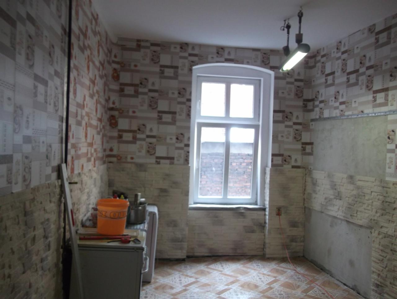 Kuchnia w trakcie remontu: położone płytki imitujące cegłę do połowy ściany, reszta ściany z tapetą, widoczne narzędzia i wiadro na kuchence, podłoga z płytek w geometryczny wzór.