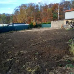 Przygotowany pod budowę teren z widoczną świeżo przekopaną ziemią, ogrodzeniem z siatki i stertą gruzu w tle.