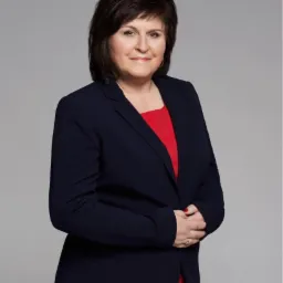 Portret kobiety w granatowej marynarce i czerwonej sukience, uśmiechająca się do kamery na szarym tle.