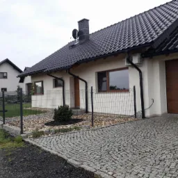 Nowoczesny dom jednorodzinny z ciemną dachówką, jasną elewacją, drewnianą bramą garażową i ogrodzeniem panelowym na kamiennej kostce brukowej.