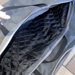 FIRMA FASTRYGA Lubow Jankowicz - Otwarta szara torba z czarną, pikowaną podszewką w romby, widoczne uchwyty i zamek błyskawiczny. Fragment dłoni w lewym górnym rogu.