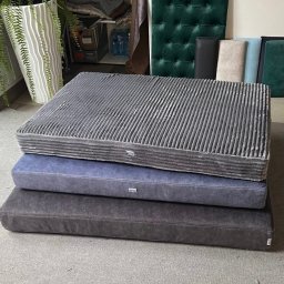 FIRMA FASTRYGA Lubow Jankowicz - Stos poduszek tapicerowanych w odcieniach szarości i błękitu, z wyraźną fakturą materiału, na tle pracowni z zielonymi panelami i roślinami.