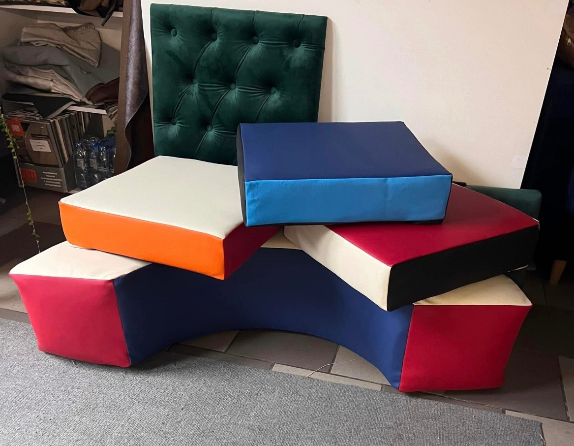 Kolorowe, geometryczne pufy i zagłówek z zielonego weluru. Pufy w odcieniach bieli, pomarańczy, czerwieni i niebieskiego, ułożone warstwowo na szarym dywanie.