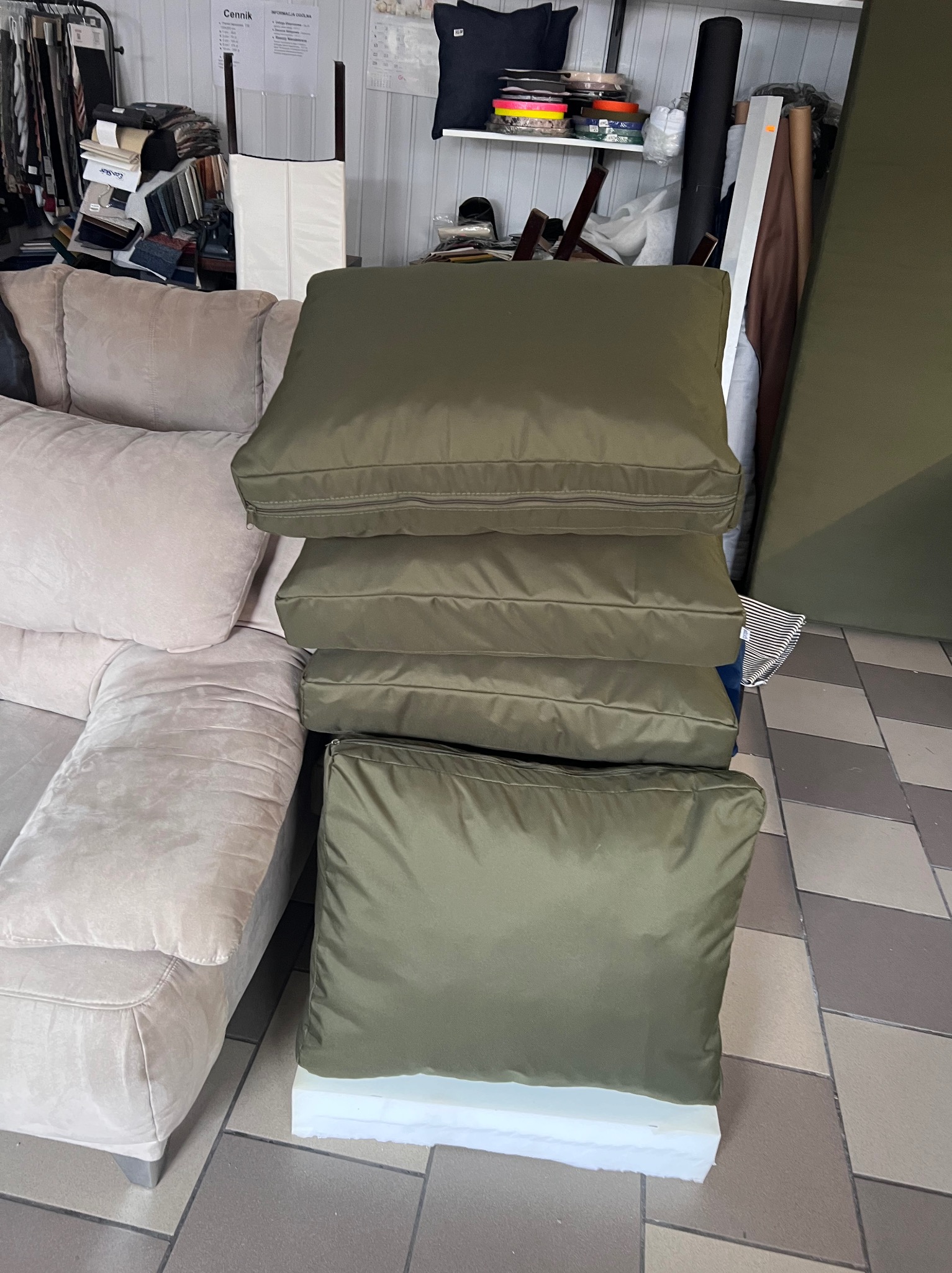 Stos poduszek w oliwkowym pokrowcu z zamkiem błyskawicznym, obok beżowej sofy. W tle widoczne materiały i akcesoria tapicerskie.