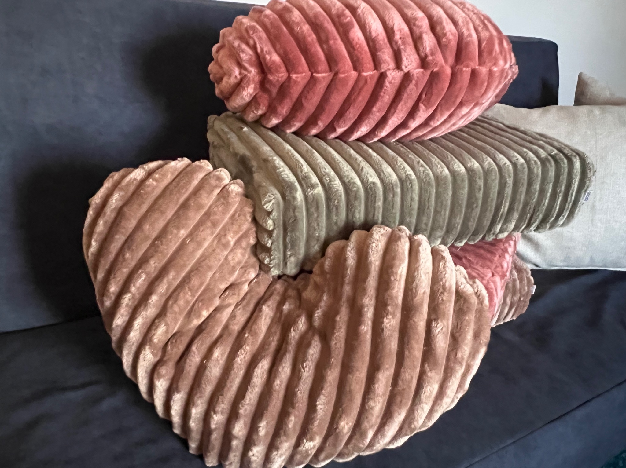 Stos poduszek dekoracyjnych z prążkowanej tkaniny w odcieniach różu i szarości na ciemnoniebieskiej sofie. Poduszka w kształcie serca na pierwszym planie.