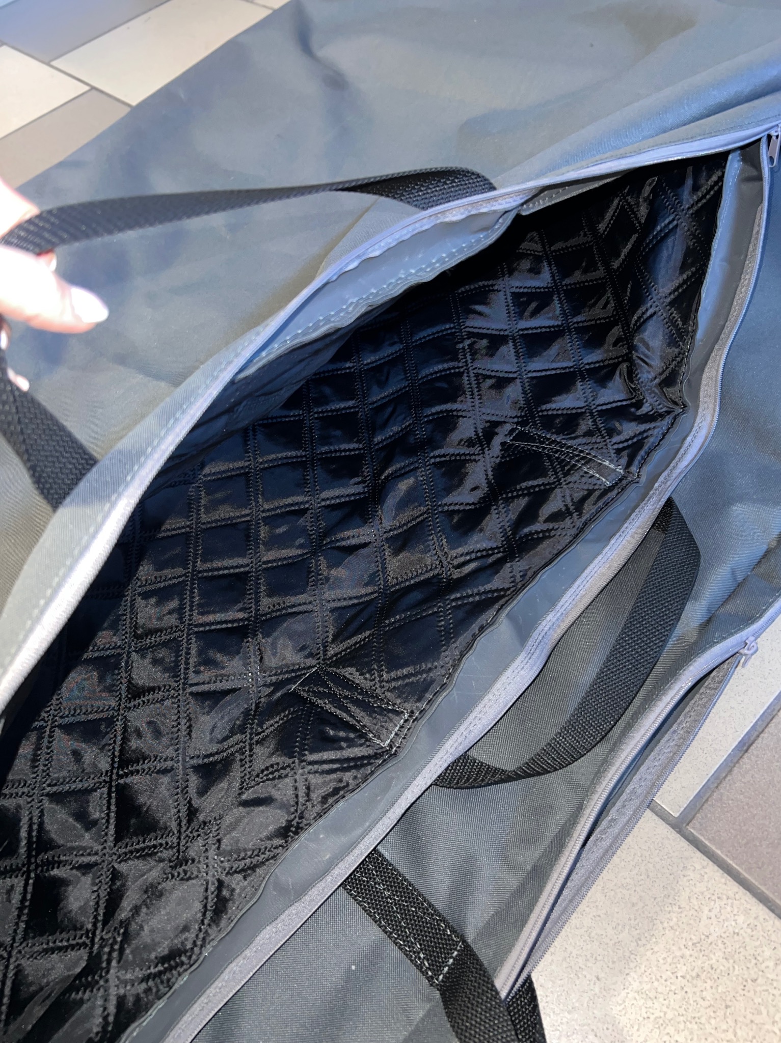 Otwarta szara torba z czarną, pikowaną podszewką w romby, widoczne uchwyty i zamek błyskawiczny. Fragment dłoni w lewym górnym rogu.