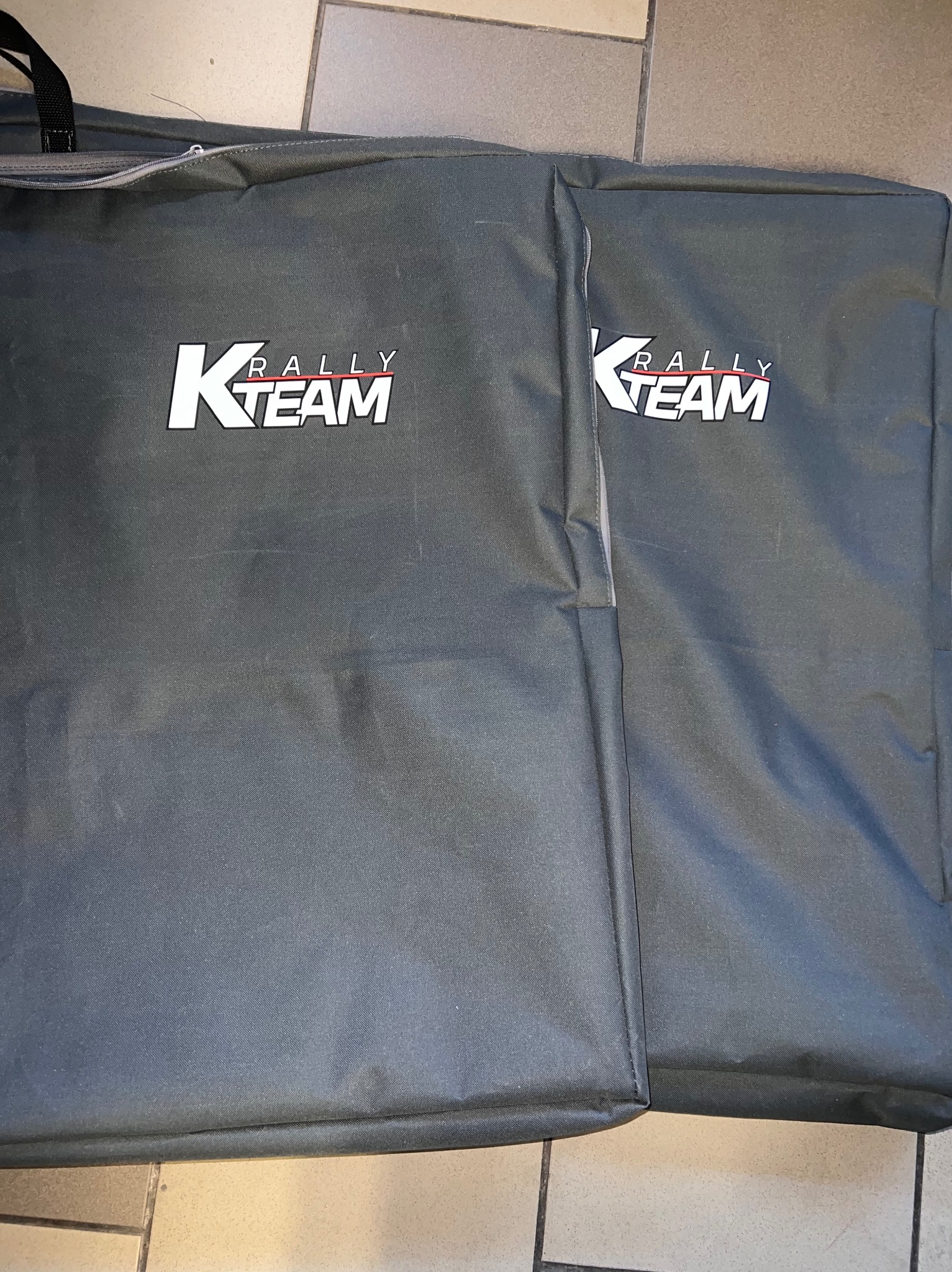 Dwa szare pokrowce z logo 'K Rally Team' leżące na podłodze z płytek. Widoczny zamek błyskawiczny i uchwyt jednego z pokrowców. Materiał wydaje się być wytrzymały.