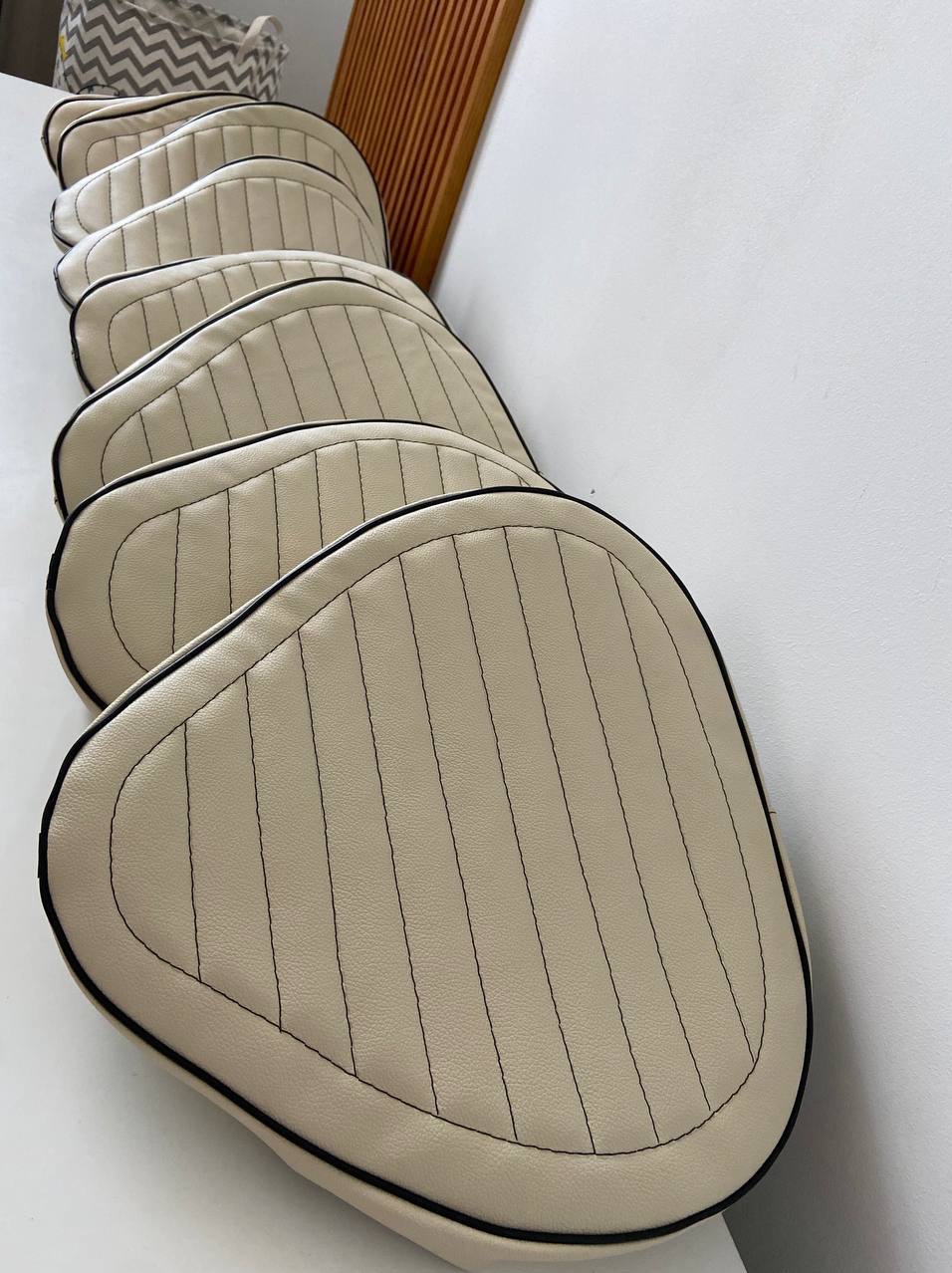 Kremowe, pikowane poduszki z czarną lamówką, ułożone rzędami na białym tle. Widoczne pionowe przeszycia i zaokrąglony kształt. Dekoracyjny element wnętrza.