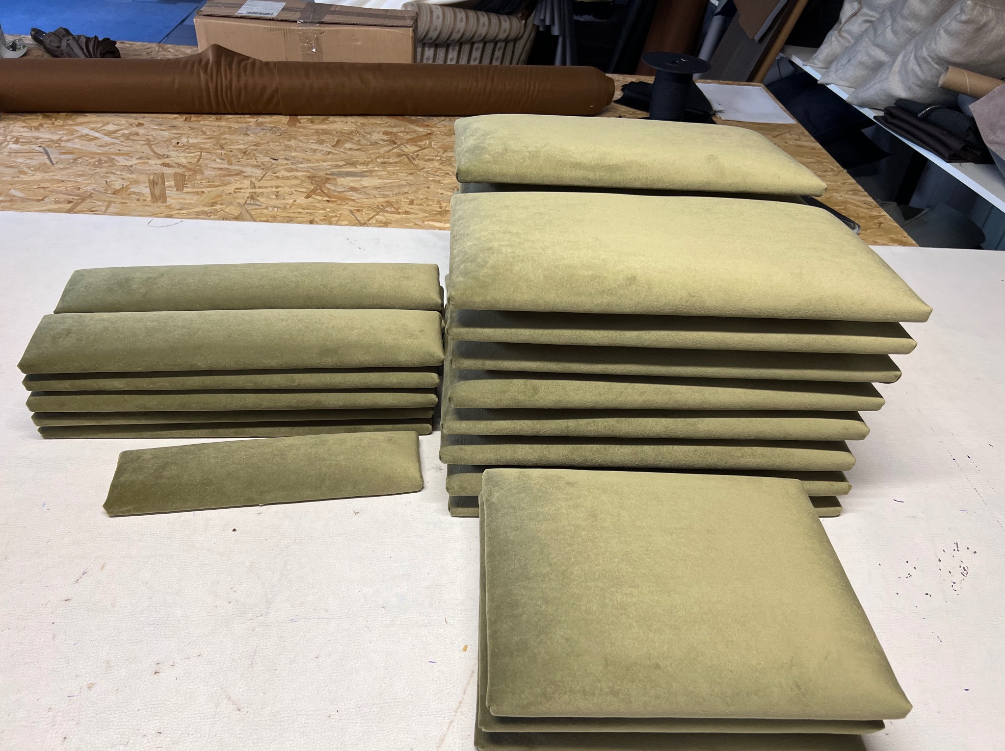 Stosy gotowych, oliwkowych poduszek i oparć krzeseł tapicerowanych welurem, ułożone na stole w warsztacie. W tle rolka brązowego materiału i akcesoria.