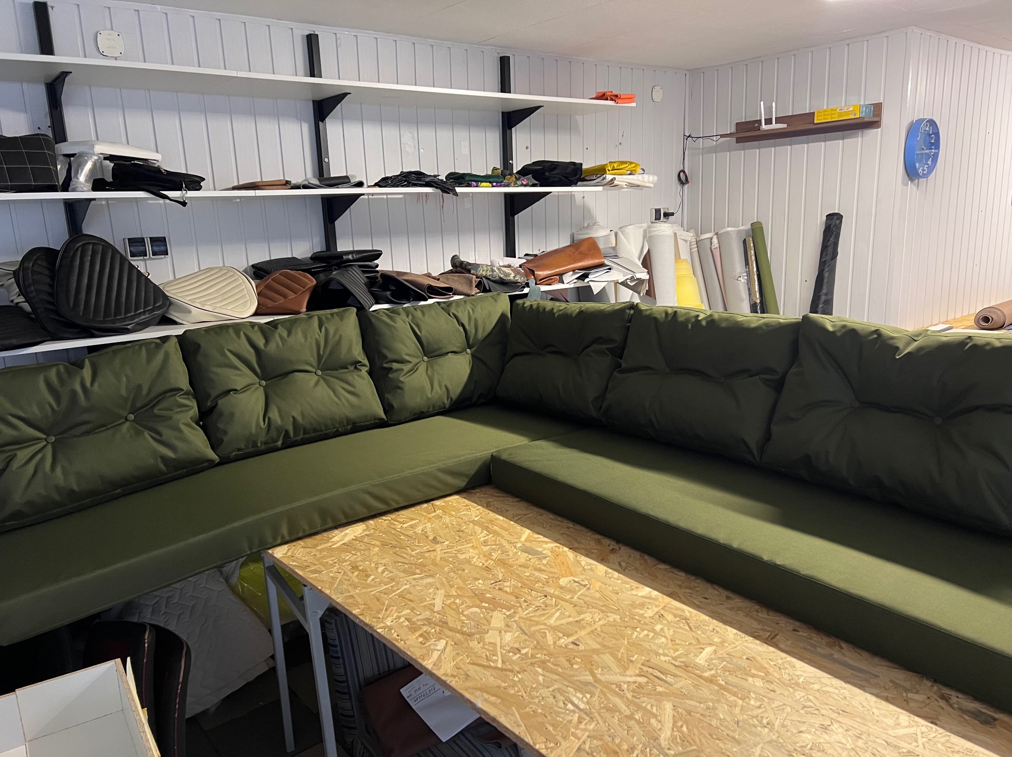 Kanciapa z zielonym narożnikiem z poduchami i blatem roboczym. Na półkach skóry i materiały. Widoczne rolki tkanin i gotowe siedziska motocyklowe.