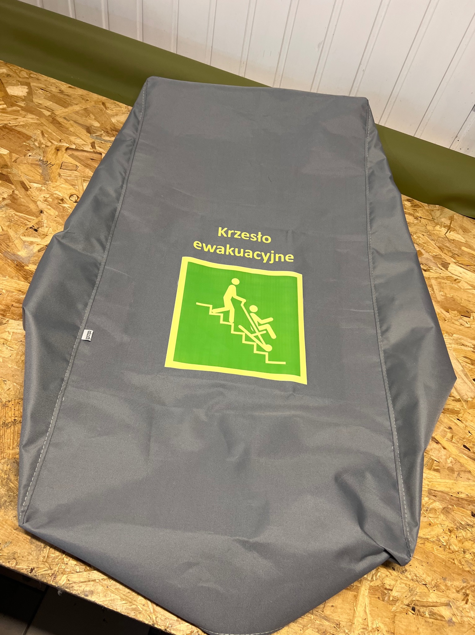 Szary pokrowiec na krzesło ewakuacyjne z zielonym symbolem i żółtym napisem, leżący na drewnianej powierzchni. Widoczne szwy i metka.