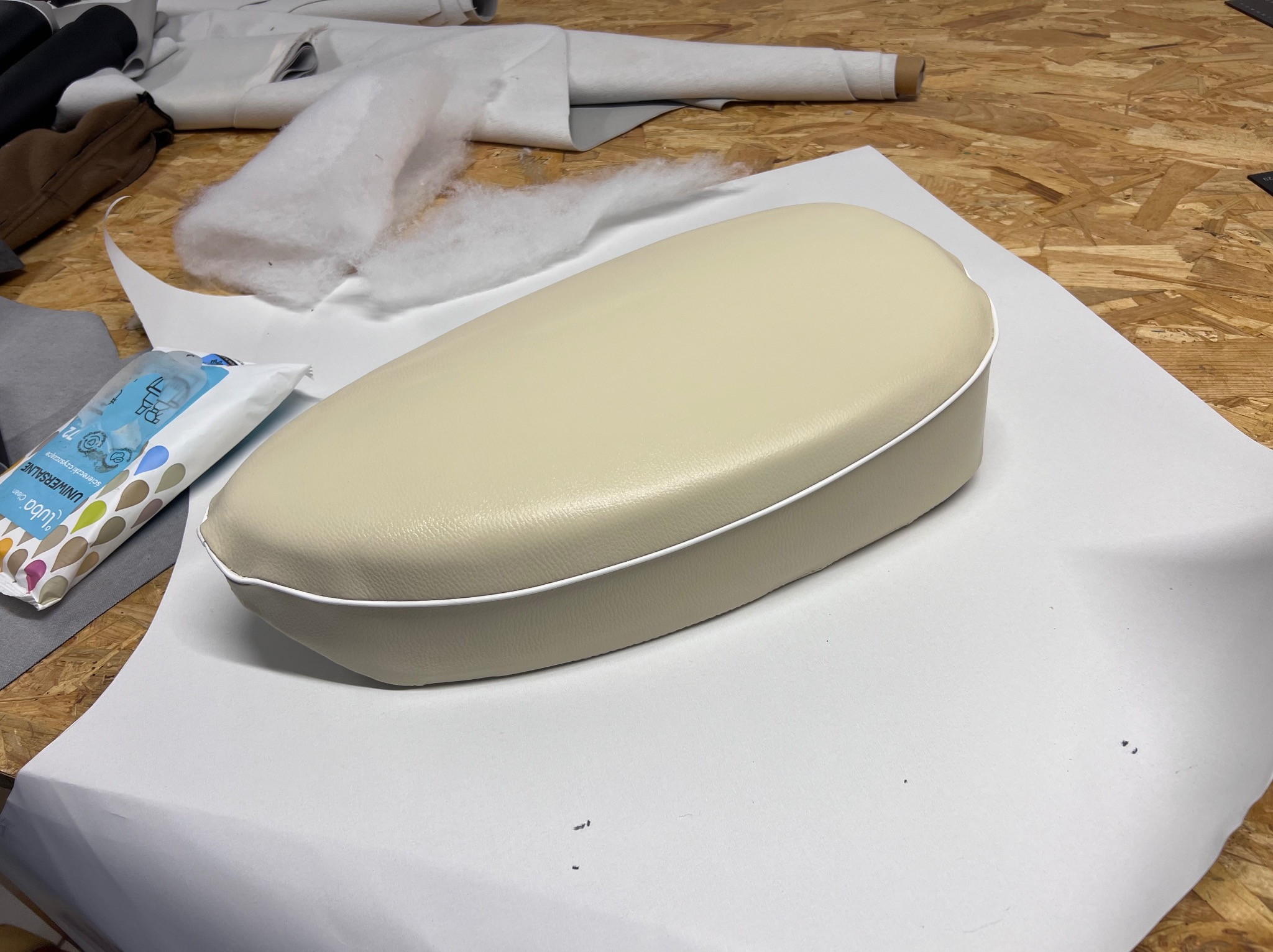 Beżowa poduszka tapicerowana z białą lamówką, leżąca na białym materiale. W tle materiały, wata i chusteczki nawilżane na drewnianym blacie.