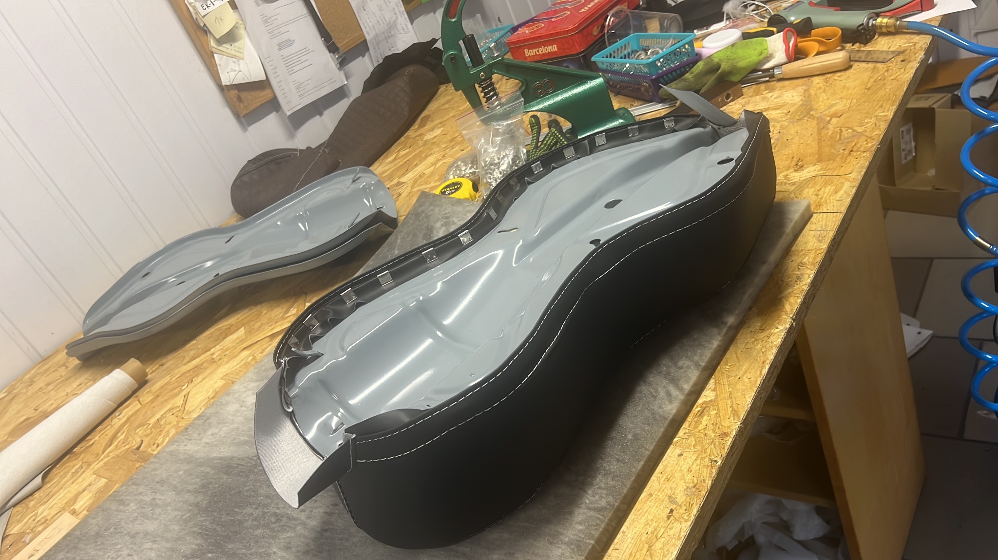 Tapicerowany element siedziska w trakcie pracy, z widocznymi narzędziami i materiałami na drewnianym stole warsztatowym. Szare wnętrze, czarne obszycie z białym szwem.