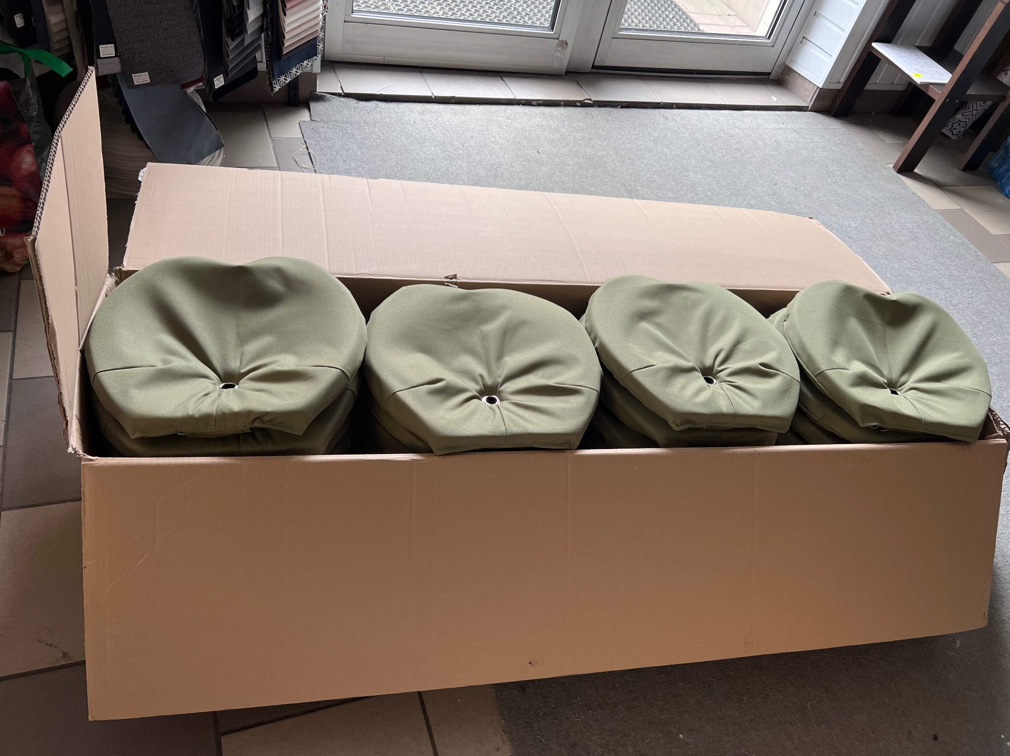 Paczka z oliwkowymi pokrowcami na krzesła/taborety, widoczne szwy i otwory na guziki. Tkaniny w tle sugerują usługi tapicerskie lub szycie na miarę.