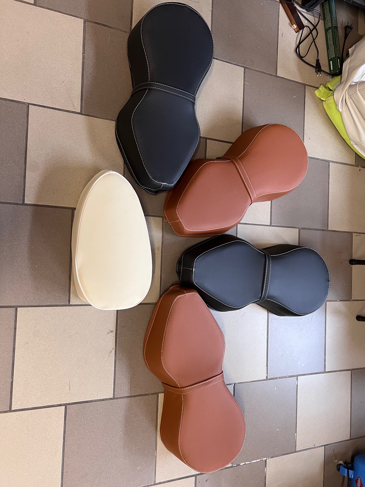 Różnokolorowe, gotowe siedziska motocyklowe z ekoskóry, z białym przeszyciem, leżące na podłodze z płytek. Widok z góry, różne kształty i odcienie brązu, beżu i czerni.