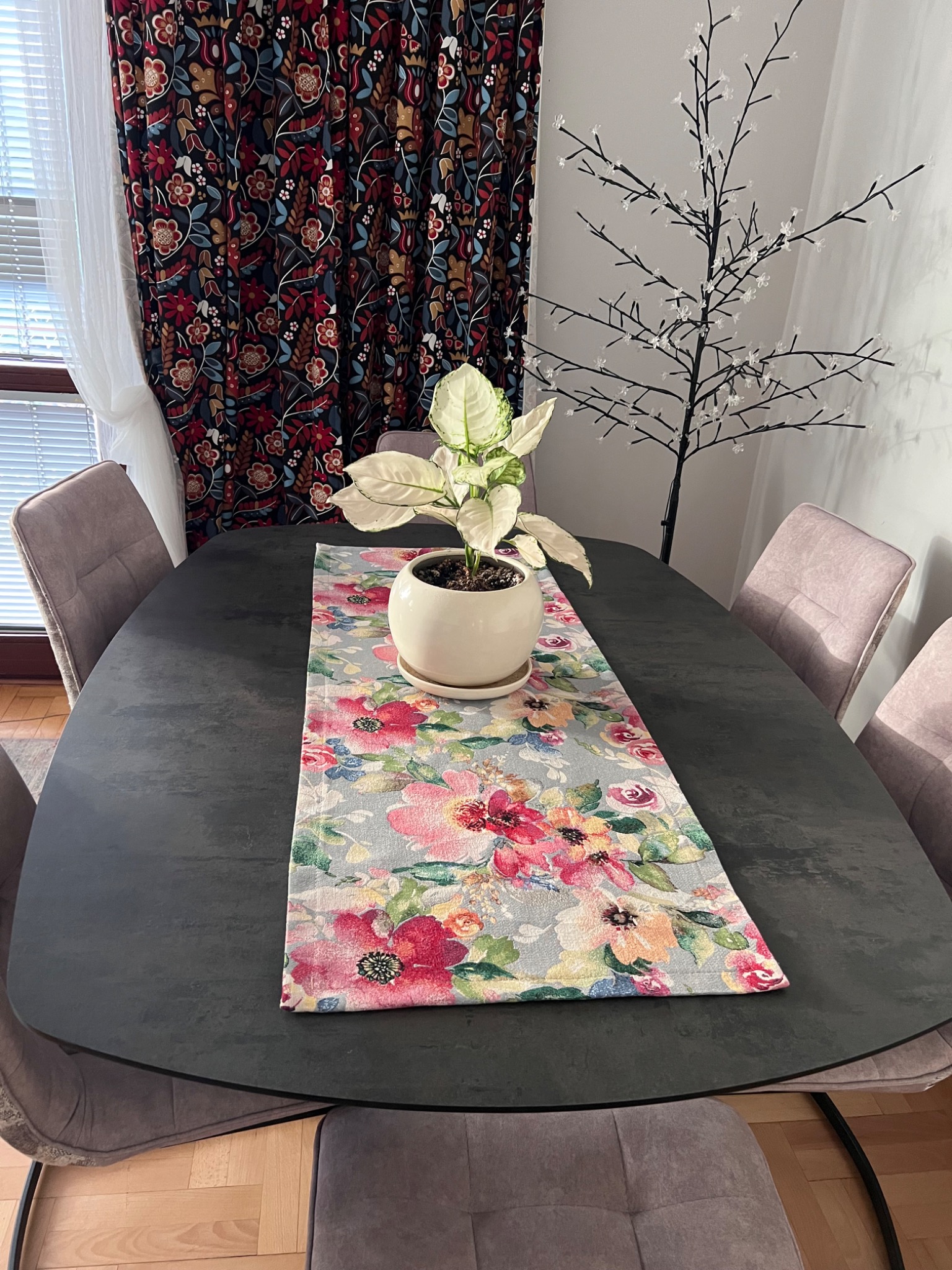 Stół z ciemnym blatem, bieżnik w kwiaty, roślina w doniczce. W tle zasłony w kwiaty i dekoracyjne drzewko. Ustawienie stołu z krzesłami w odcieniach fioletu.