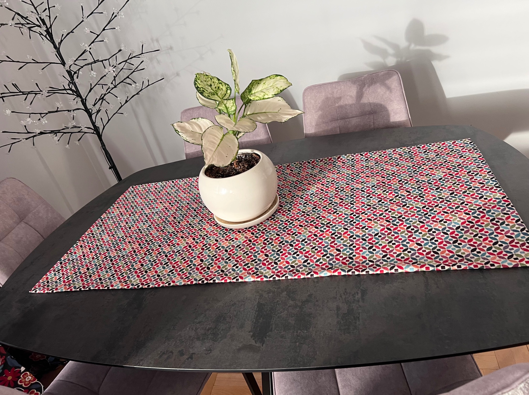 Stół z ciemnym blatem, nakryty wzorzystym bieżnikiem w odcieniach czerwieni, błękitu i zieleni. Na nim doniczka z rośliną. W tle krzesła z fioletową tapicerką.
