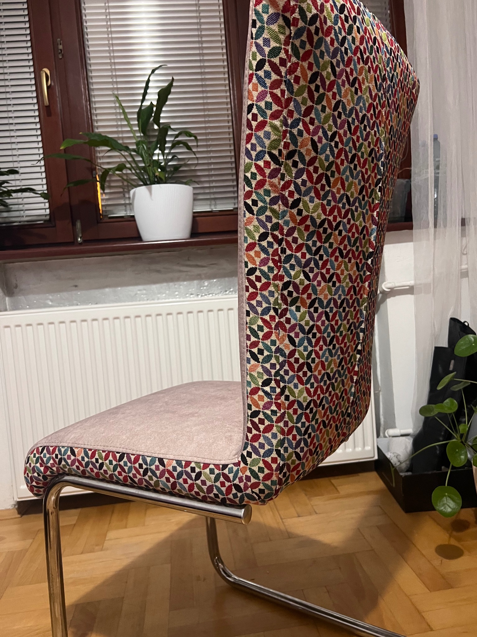 Krzesło po renowacji z nową tapicerką w kolorowy wzór, na metalowych nogach. W tle okno z żaluzjami i roślina doniczkowa. Widok z boku, na podłodze parkiet.