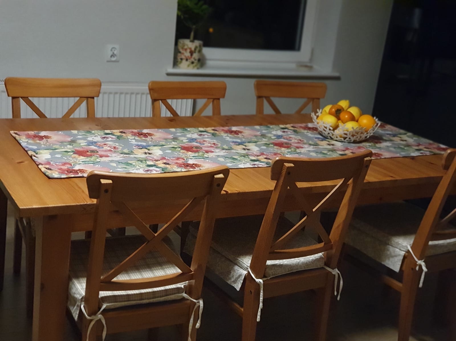 Drewniany stół z bieżnikiem w kwiaty i krzesłami z poduszkami. Na stole misa z owocami, w tle roślina w doniczce na parapecie okna.