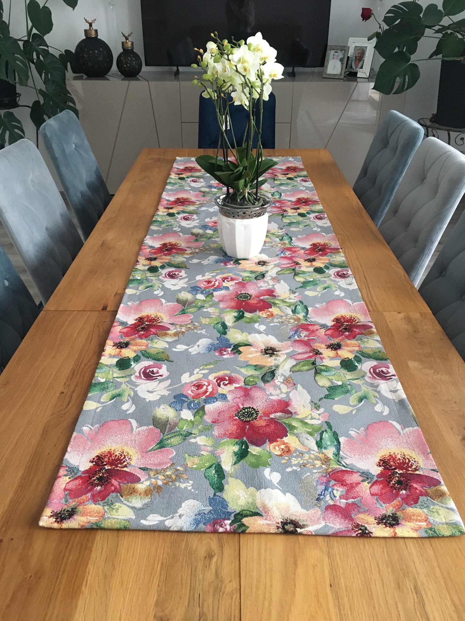 Stół z drewnianym blatem, na którym leży długi, kwiecisty bieżnik. W tle widoczne krzesła z szarym obiciem i rośliny doniczkowe. Na stole stoi doniczka z białymi kwiatami.