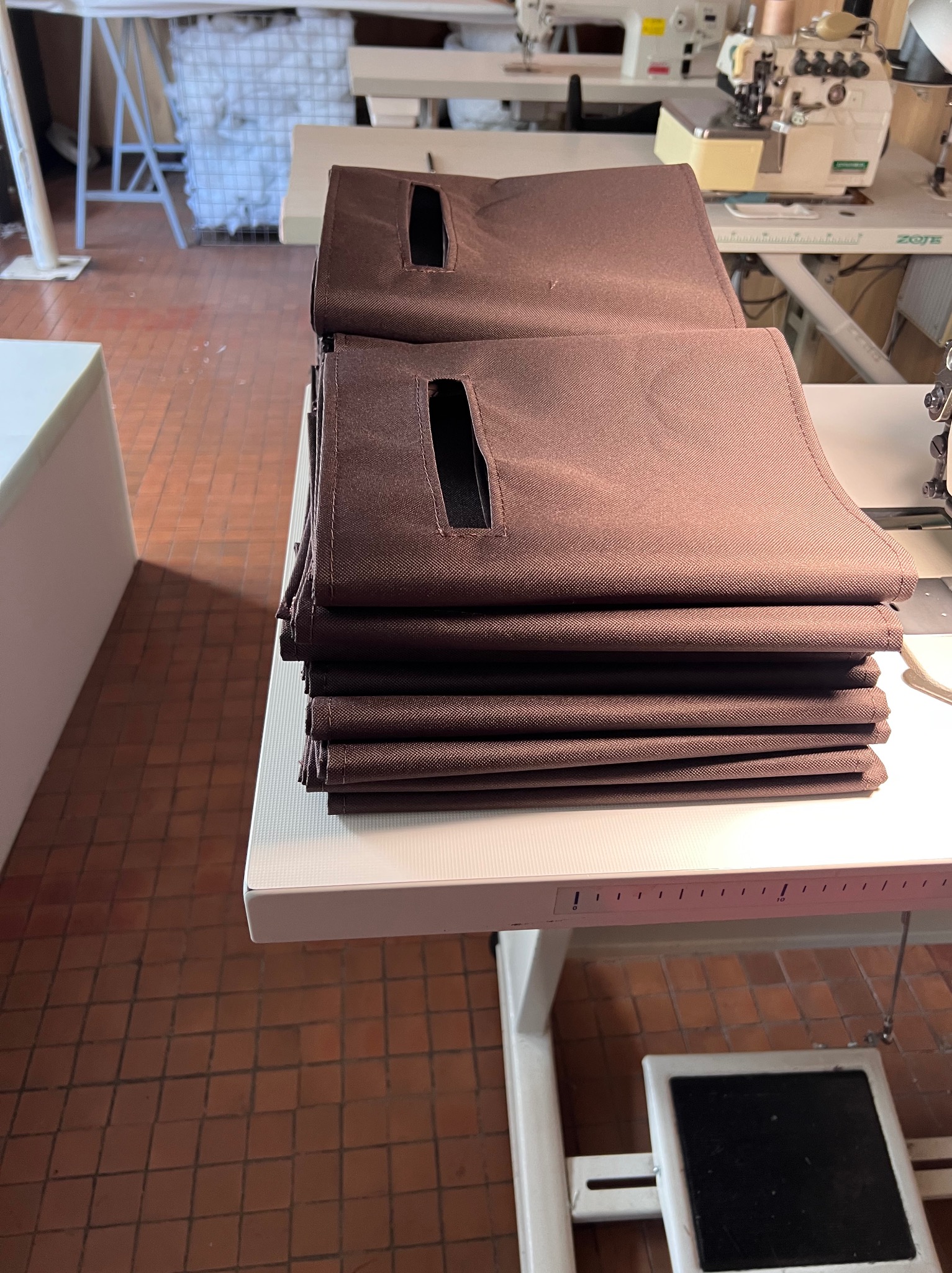 Stos brązowych pokrowców z uchwytami na stole szwalniczym, w tle maszyny do szycia i materiały. Widoczne detale wykończenia i faktura tkaniny.