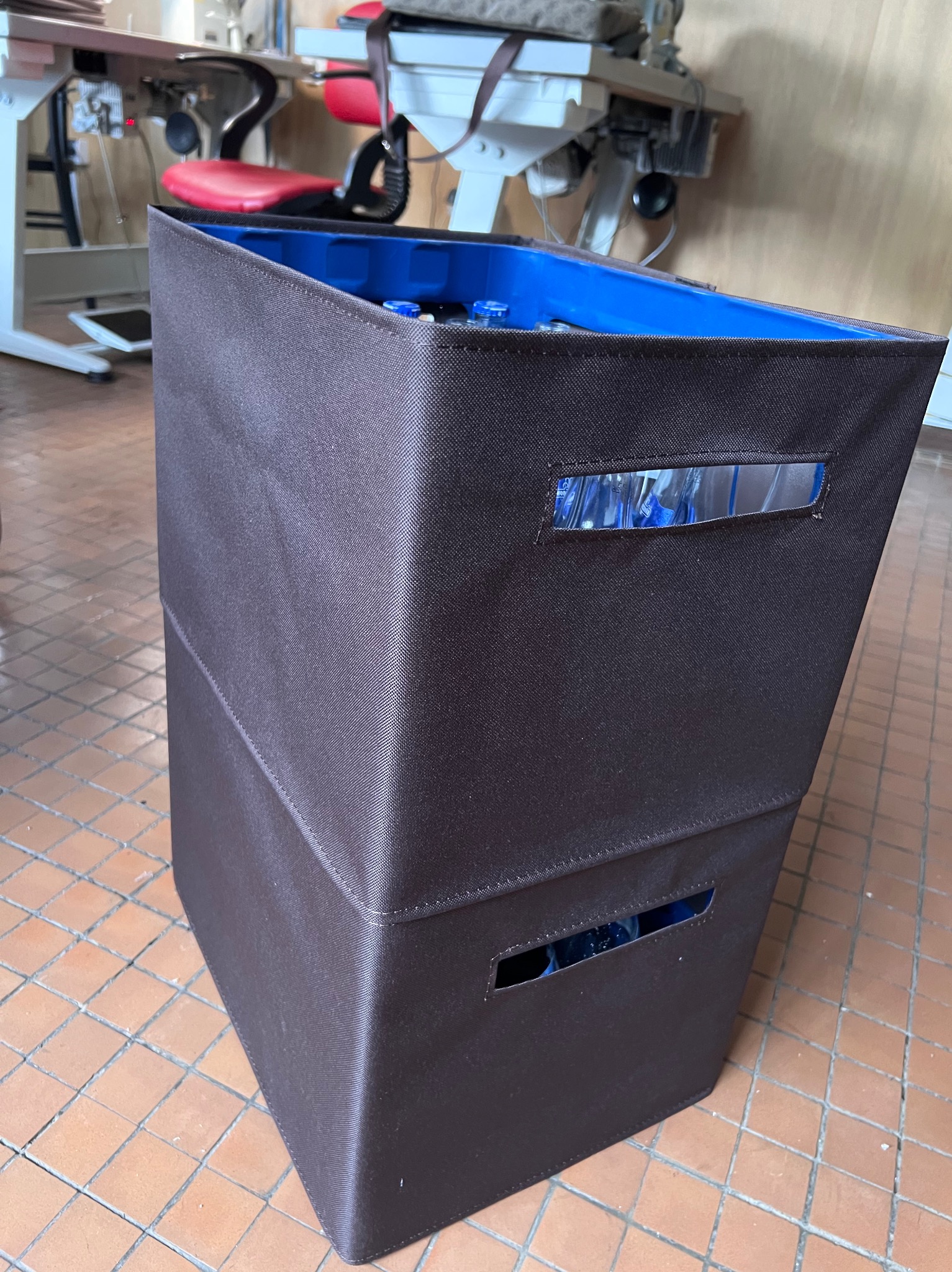 Dwuwarstwowy, brązowy pojemnik z tkaniny z niebieskim wnętrzem, stojący na podłodze z terakoty. Widoczne okienka z zawartością w środku.