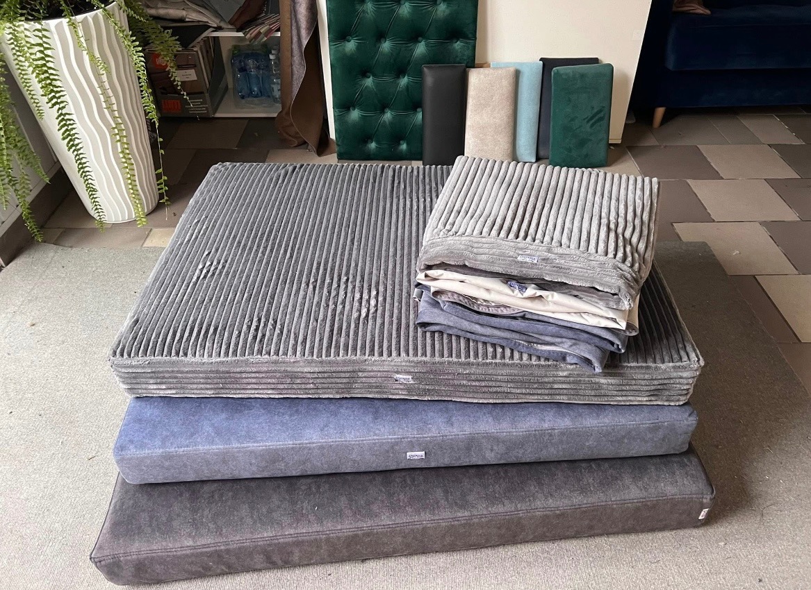 Stos poduszek tapicerowanych w odcieniach szarości i błękitu, obok pionowe panele tapicerowane w różnych kolorach i fakturach. Widoczny fragment sofy w tle.