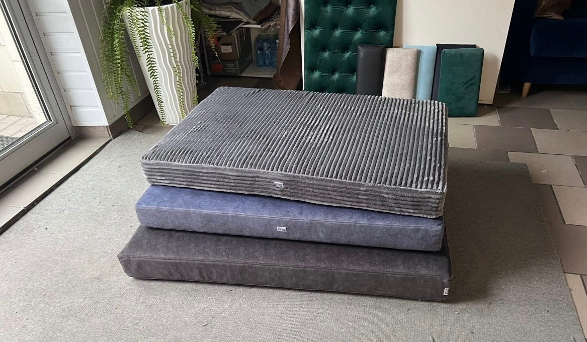 Stos poduszek tapicerowanych w odcieniach szarości i błękitu, z wyraźną fakturą materiału, na tle pracowni z zielonymi panelami i roślinami.