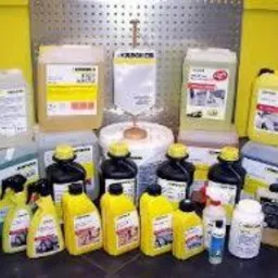 Zestaw różnorodnych środków czyszczących w plastikowych pojemnikach i butelkach, głównie w kolorach żółtym, białym i czarnym, ustawionych na podłodze na tle metalowej siatki.