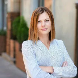 Portret kobiety z Płocka, z rudymi włosami do ramion, ubranej w bluzkę w delikatny wzór, na rozmytym tle miejskim z zielenią. Uśmiech, skrzyżowane ręce.