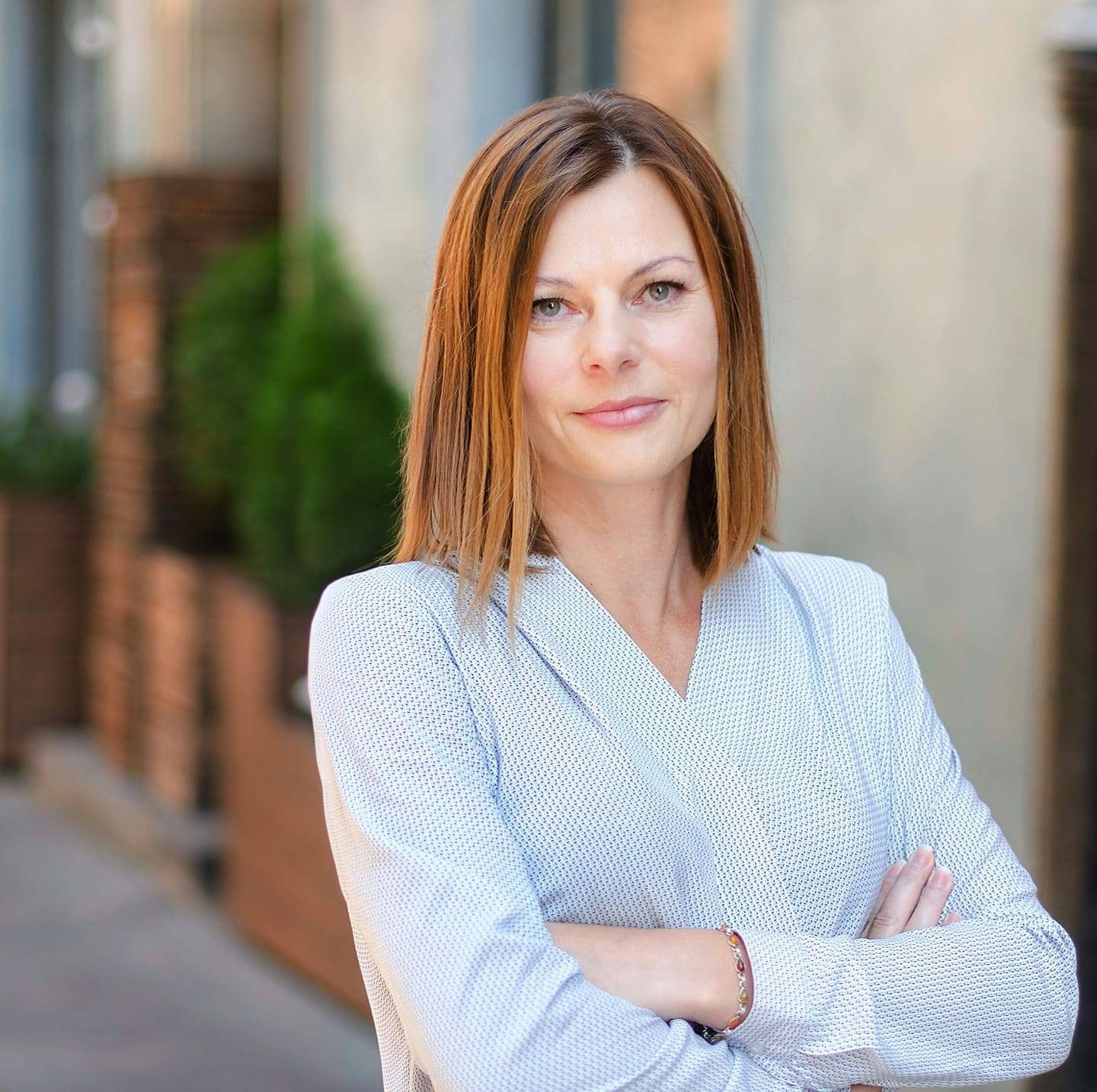 Portret kobiety z Płocka, z rudymi włosami do ramion, ubranej w bluzkę w delikatny wzór, na rozmytym tle miejskim z zielenią. Uśmiech, skrzyżowane ręce.