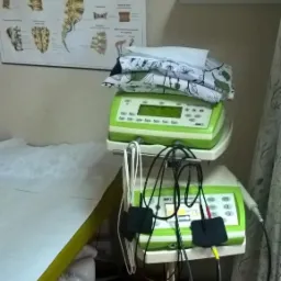 Stół do zabiegów z zielonymi urządzeniami do elektroterapii na wózku, obok plakat z budową kręgosłupa i półka z akcesoriami.