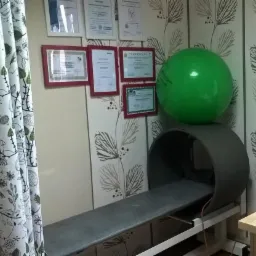 Gabinet z rollmasażem, piłką do ćwiczeń, dyplomami w ramkach i zasłoną w kwiaty.