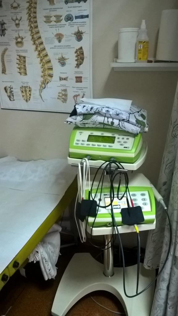Stół do zabiegów z zielonymi urządzeniami do elektroterapii na wózku, obok plakat z budową kręgosłupa i półka z akcesoriami.