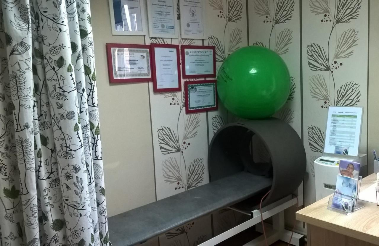 Gabinet z rollmasażem, piłką do ćwiczeń, dyplomami w ramkach i zasłoną w kwiaty.