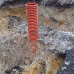 Pomarańczowa, karbowana rura kanalizacyjna wystaje z wykopu w ziemi o różnej strukturze i kolorystyce, widoczny fragment taśmy ostrzegawczej oraz narzędzia.