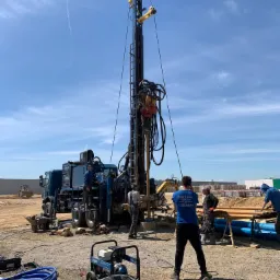 Wiercenie studni głębinowej w toku: ekipa w niebieskich koszulkach nadzoruje pracę wiertnicy na podwoziu ciężarówki, widoczne rury, agregat prądotwórczy i sprzęt hydrauliczny na placu budowy.