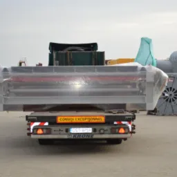 Transport niskopodwoziowy przewozi zabezpieczony folią element konstrukcyjny o metalowej ramie, widoczne tablice ostrzegawcze 'Convoi Exceptionnel' oraz bębny kablowe w tle.