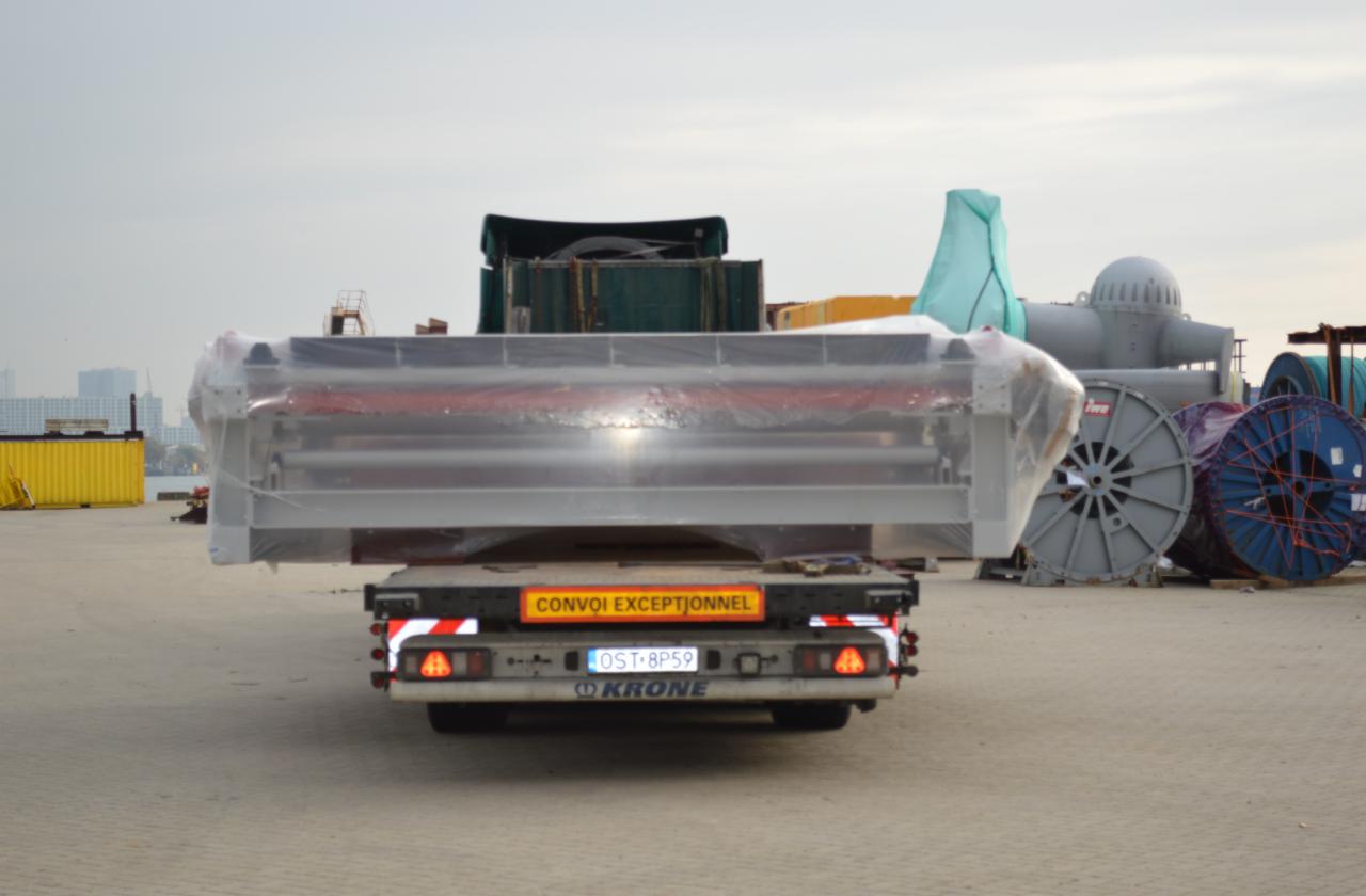 Transport niskopodwoziowy przewozi zabezpieczony folią element konstrukcyjny o metalowej ramie, widoczne tablice ostrzegawcze 'Convoi Exceptionnel' oraz bębny kablowe w tle.