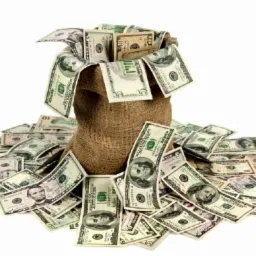 Jutowy worek wypełniony i otoczony plikiem banknotów dolarowych o różnych nominałach na białym tle.