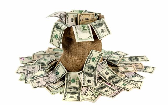 Jutowy worek wypełniony i otoczony plikiem banknotów dolarowych o różnych nominałach na białym tle.