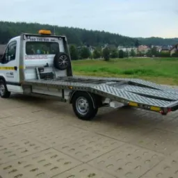 Biała laweta z zapasowym kołem na tle zielonego krajobrazu, widoczna na betonowej nawierzchni.