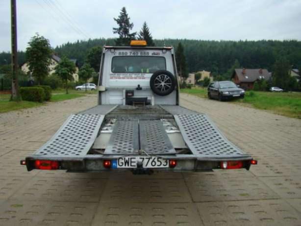 Widok z tyłu na lawetę z rozłożonymi najazdami na betonowej nawierzchni, w tle zalesiony teren i zabudowania.