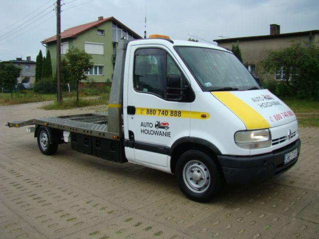 Biała laweta Renault Master z żółtymi pasami i numerem telefonu, zaparkowana na brukowanej drodze przed budynkami.