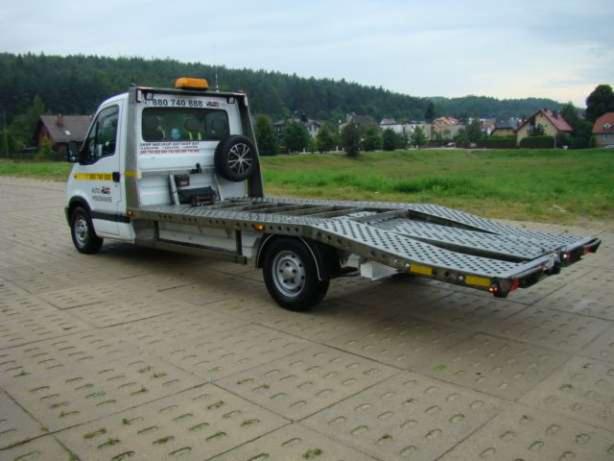 Biała laweta z zapasowym kołem na tle zielonego krajobrazu, widoczna na betonowej nawierzchni.