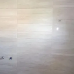 Ściana wyłożona beżowymi płytkami imitującymi marmur, z widocznymi otworami na armaturę w łazience w trakcie prac wykończeniowych.